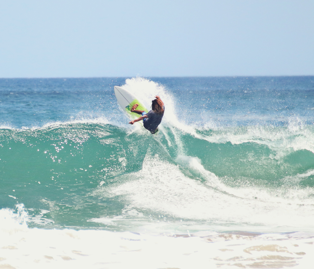 A person with long hair performs a maneuver on a surfboard, catching air above the crest of a large ocean wave. The sea is a vibrant blue, and white foam is visible where the wave breaks. The scene is set under a clear sky.