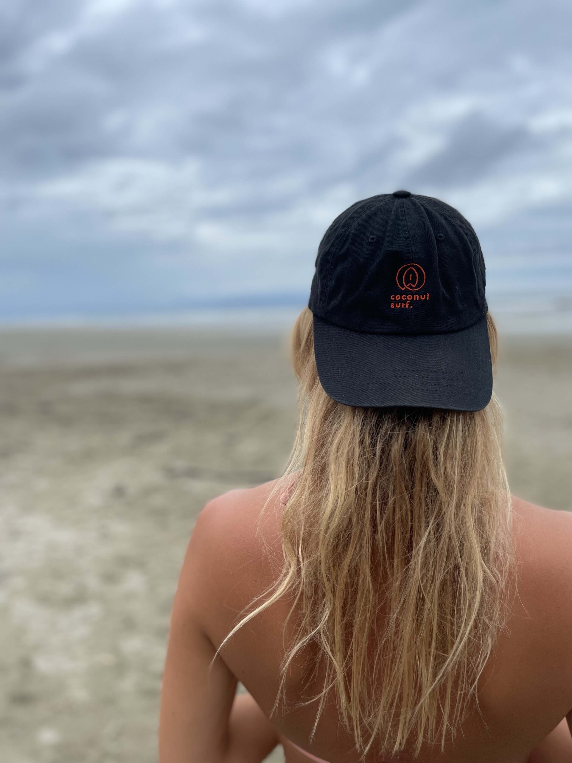 A person with long blond hair is facing a cloudy beach and wearing a black cap with a orange logo and the text "coconut surf." The sandy beach and overcast sky stretch into the background. The person is seen from behind.