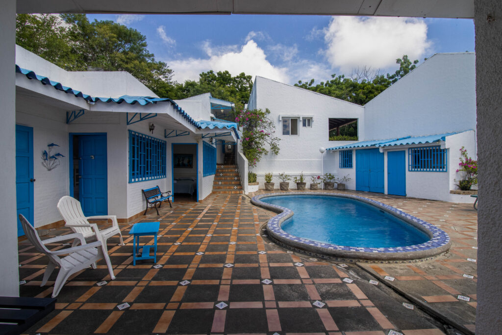 A courtyard with a geometric tiled floor featuring an irregularly shaped swimming pool surrounded by white buildings with blue doors and windows. There are chairs and a table near the pool, plants along the wall, and a staircase leading to an upper level.