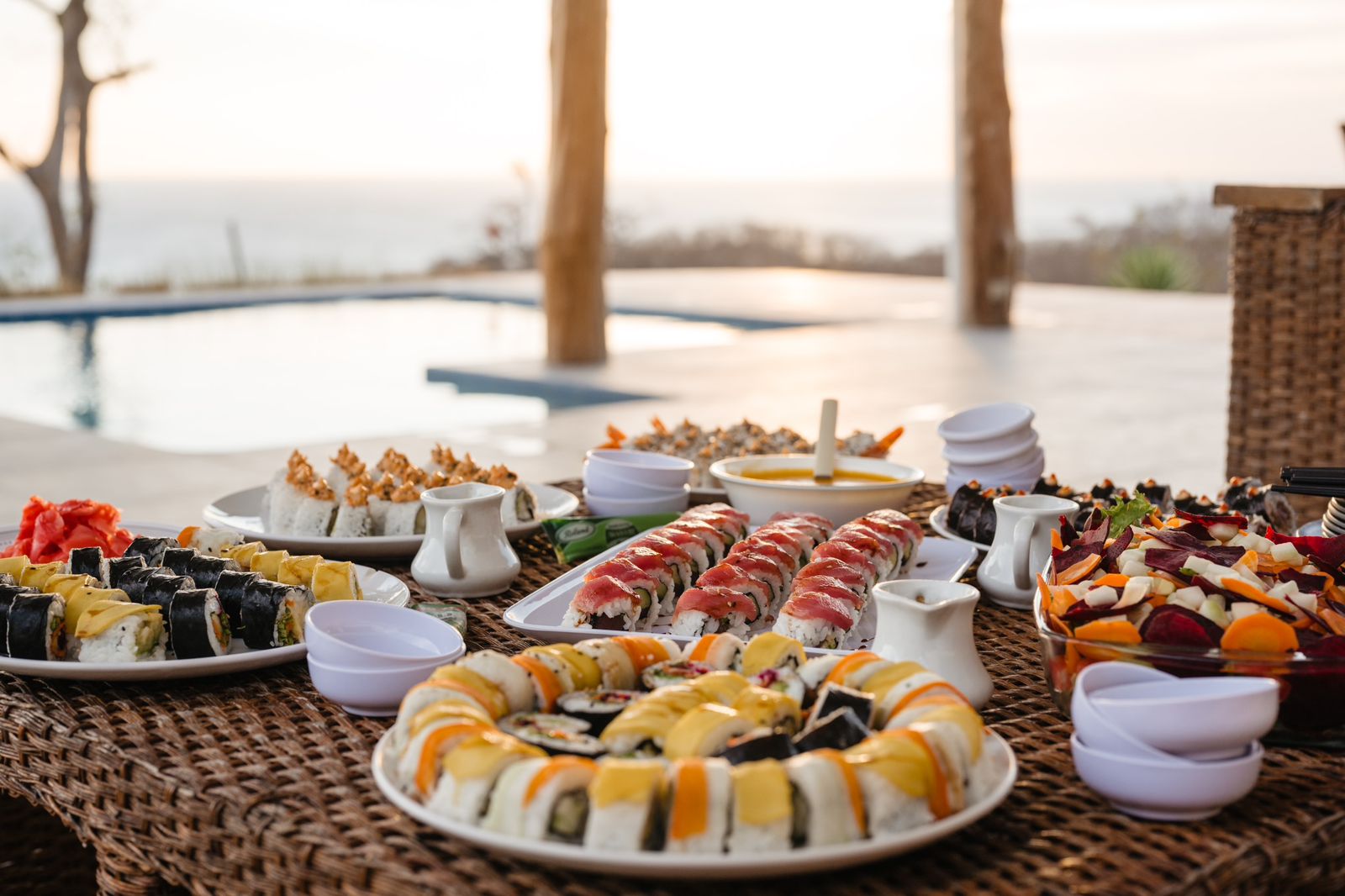 A variety of colorful sushi rolls and dishes are arranged on a wicker table outdoors by a poolside, offering unique dining experiences with dipping sauces and a beautiful sunset in the background.